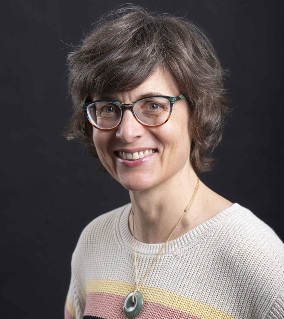 Portretfoto van pianodocente en muzikante Sharon Stewart Studioportreit van een persoon met bril, kort haar en gestreepte trui tegen een donkere achtergrond